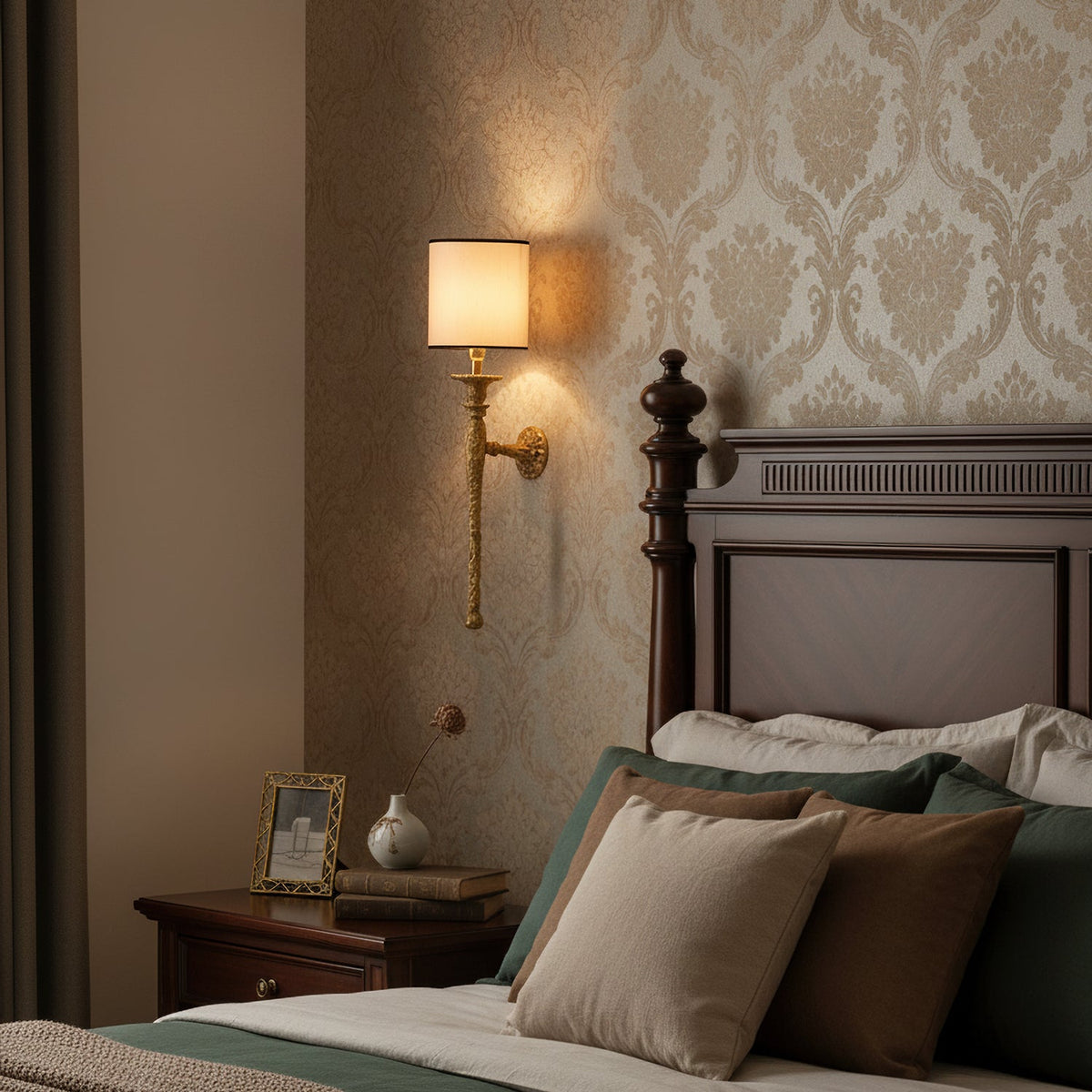 A lit beige-shaded Clérachic wall light hangs on damask wallpaper above a bedside table and a dark wooden bed with green and taupe cushions.
