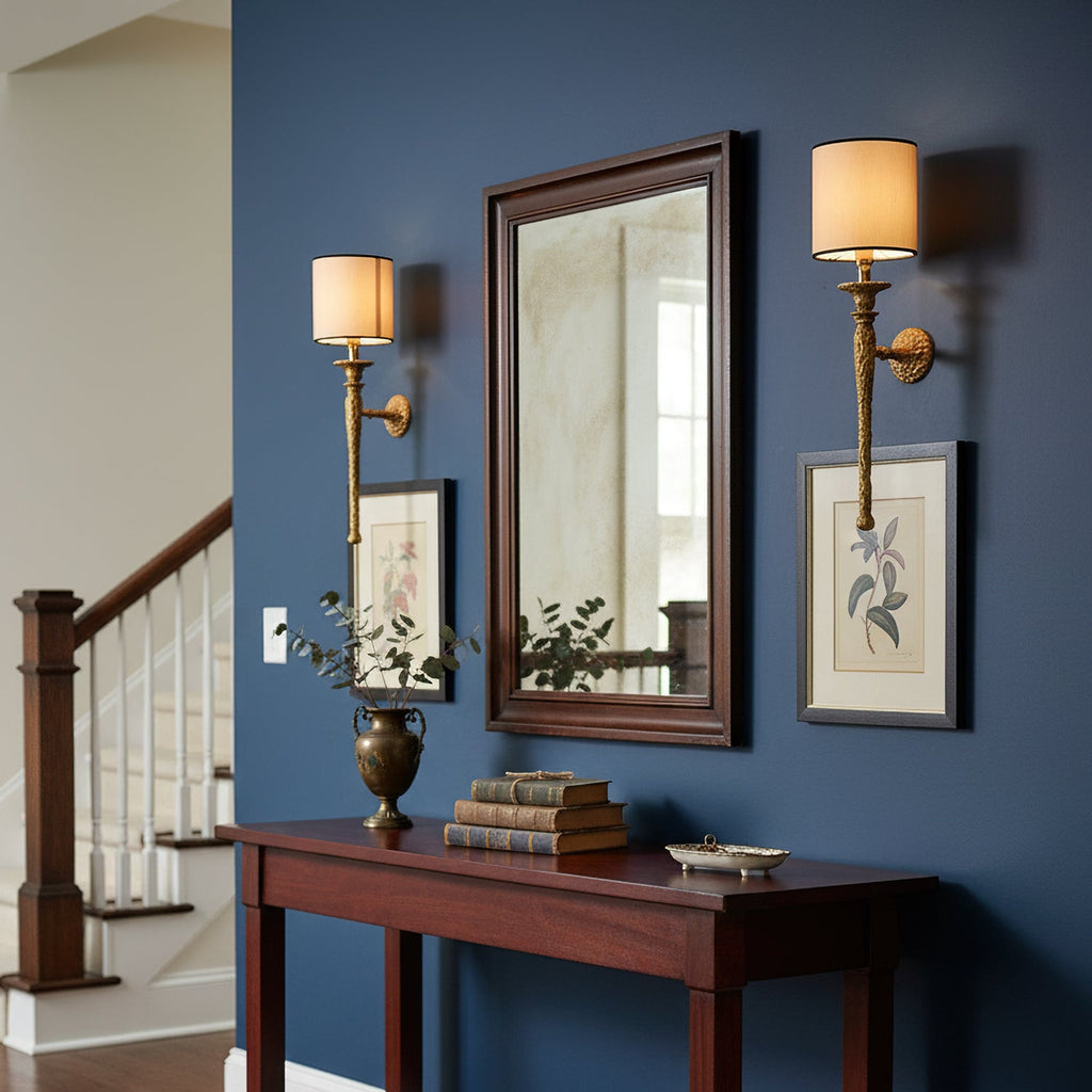 Two Clérachic brass wall lights frame a dark-wood mirror on a navy wall, above a hall console and near a staircase.