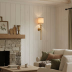 This Clérachic wall light glows on a white panelled wall beside a stone fireplace and a cream sofa with green cushions.