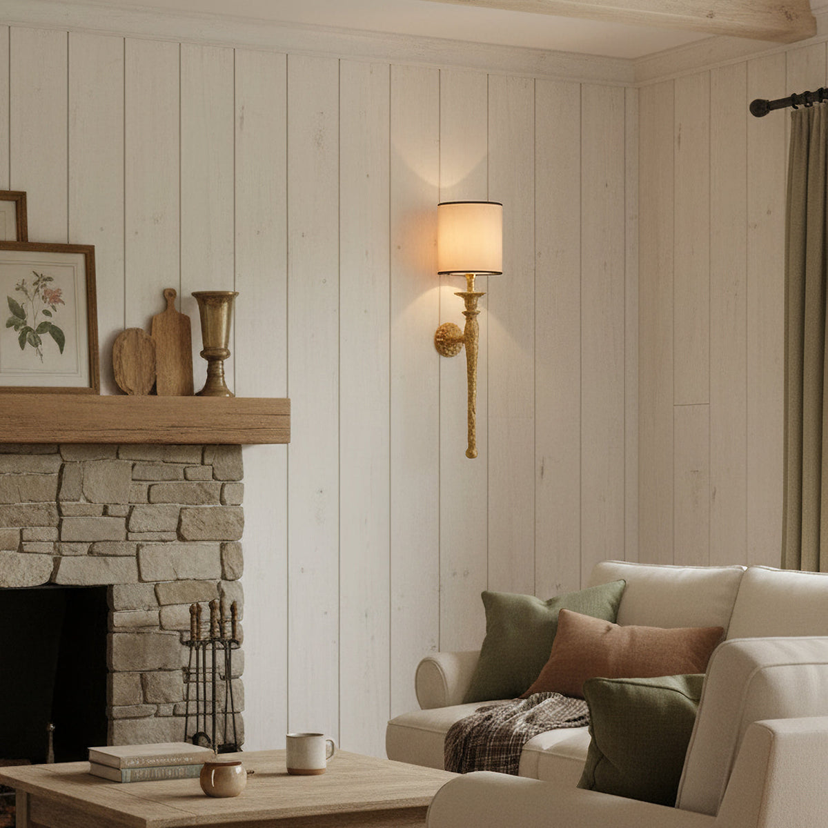 This Clérachic wall light glows on a white panelled wall beside a stone fireplace and a cream sofa with green cushions.