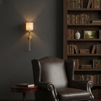 Small Clérachic brass wall light above a dark leather armchair, with a bookcase of old books and a dark grey wall behind.