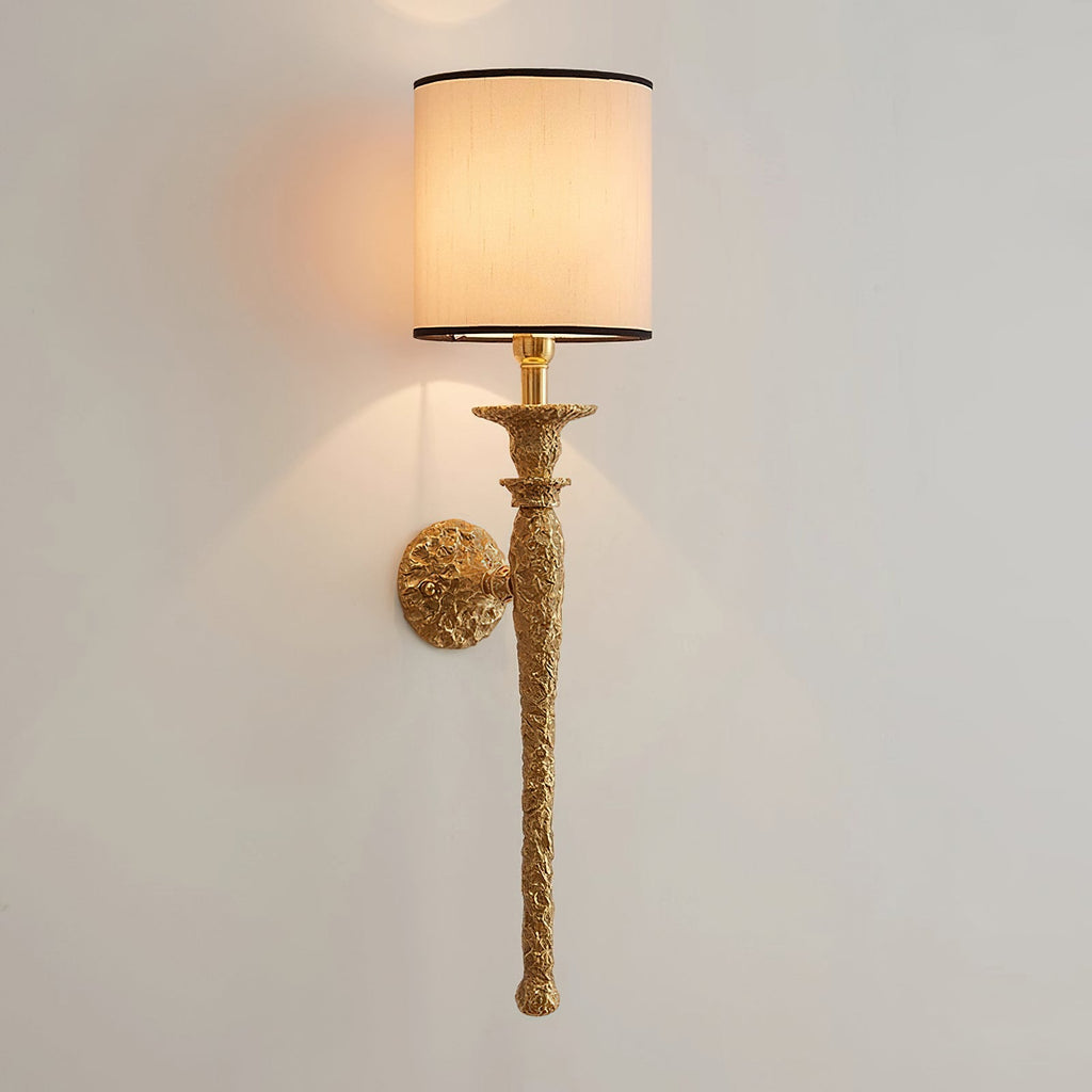 The lit beige shade and hammered antique brass stem of this Clérachic wall light on a pale wall, with light washing up and down.