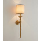 Front view of this Clérachic wall light on a light wall, its beige shade glowing above a textured brass torch-shaped stem.