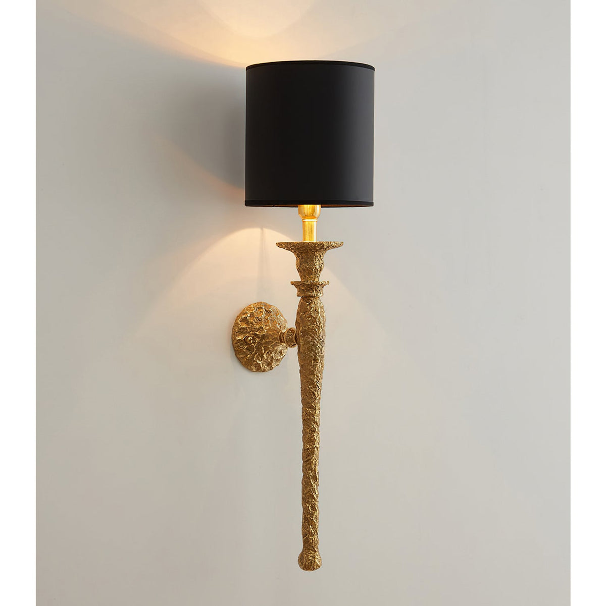 The black-shaded version of this Clérachic wall light, lit on a pale wall above its rough-cast antique brass stem.