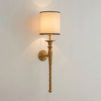 This Clérachic wall light with its beige shade lit, seen from a distance on a pale wall, showing the full slim brass stem.