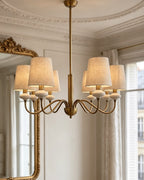 Gold Clérachic chandelier seen from slightly below, with six linen shades and looped arms against a white panelled wall.