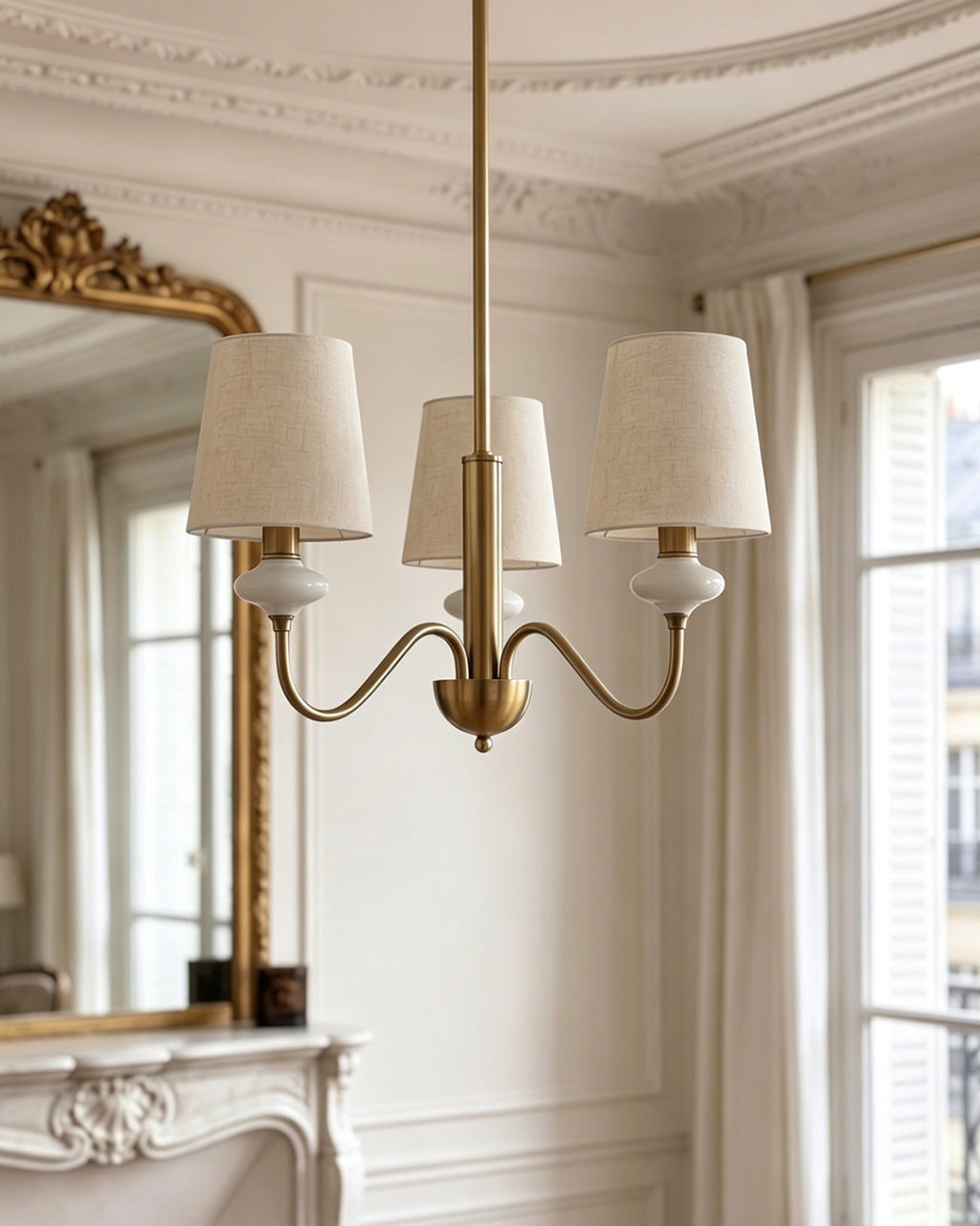 A three-light version of this Clérachic chandelier in gold, with three linen shades on curved arms in a bright, white-panelled room.
