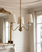 A three-light version of this Clérachic chandelier in gold, with three linen shades on curved arms in a bright, white-panelled room.