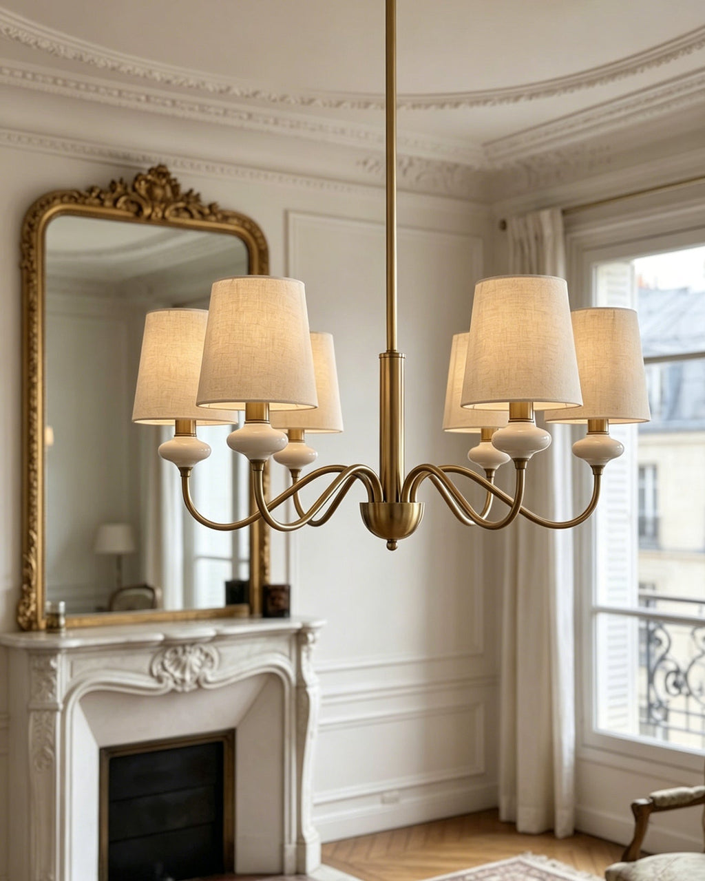 Gold Clérachic chandelier hung low over a herringbone floor, with a marble fireplace, a gilt mirror and a balcony window behind.