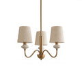 Gold three-light Clérachic chandelier with linen shades, ceramic necks and curved arms, on a plain white backdrop.