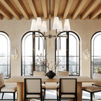 Above a long oak dining table with upholstered chairs, a Clérachic six-light chandelier hangs beneath a timber-beamed ceiling and arched windows.