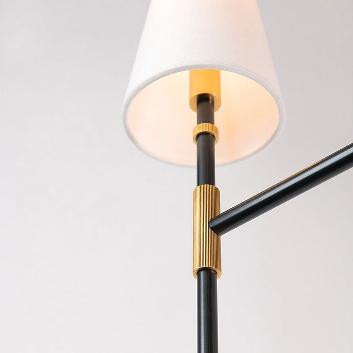 A white linen shade above a ribbed brass collar on a black arm of this Clérachic chandelier, seen from below against a pale grey wall.