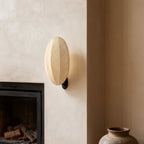 Beside a fireplace and a clay pot, this Clérachic wall light shows an oval linen shade on a small black mount against a plaster wall.