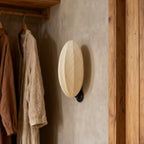 Against a plaster wall next to hanging linen shirts and a timber door frame, this Clérachic wall light shows its ribbed oval shade.