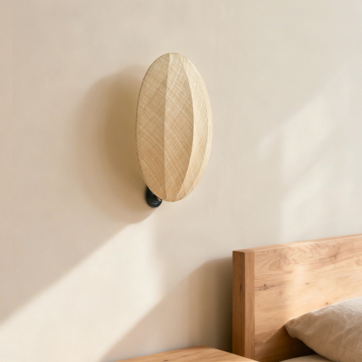 Above a wooden bed head, a Clérachic wall light throws a soft shadow on a sunlit plaster wall, its linen shade angled up.