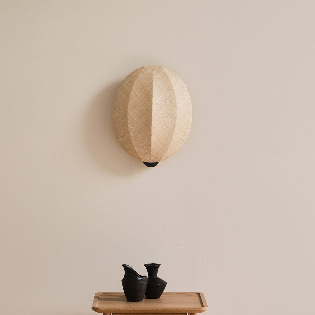 Over a small tray with two black vases, this Clérachic wall light shows its rounded linen shade with a vertical seam on a cream wall.