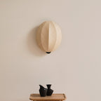 Over a small tray with two black vases, this Clérachic wall light shows its rounded linen shade with a vertical seam on a cream wall.