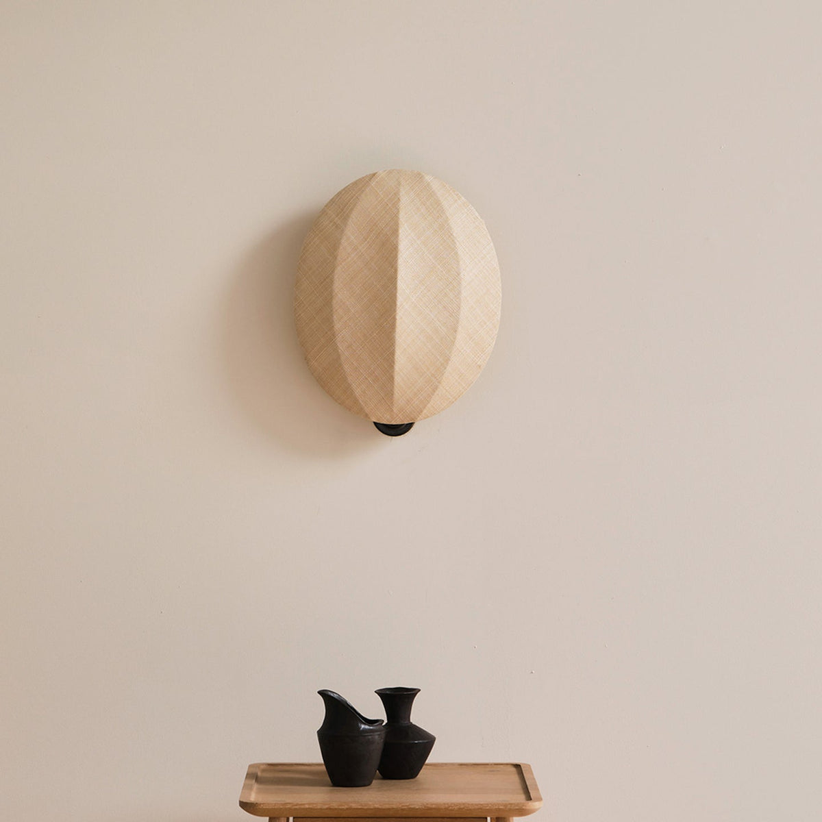 Over a small tray with two black vases, this Clérachic wall light shows its rounded linen shade with a vertical seam on a cream wall.