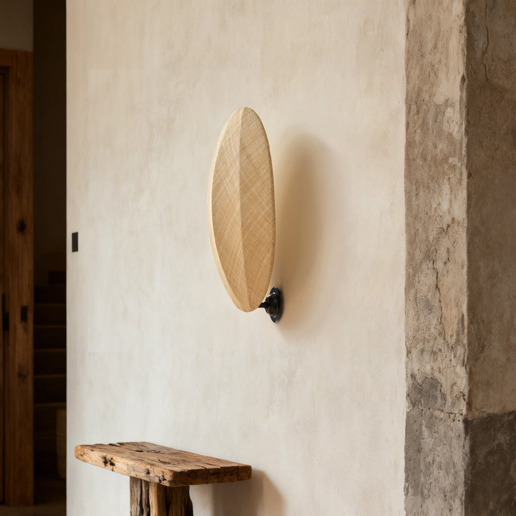 On a rough plaster wall above a rustic wooden bench, this Clérachic wall light shows its slim oval linen shade on a black mount.