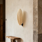 On a rough plaster wall above a rustic wooden bench, this Clérachic wall light shows its slim oval linen shade on a black mount.