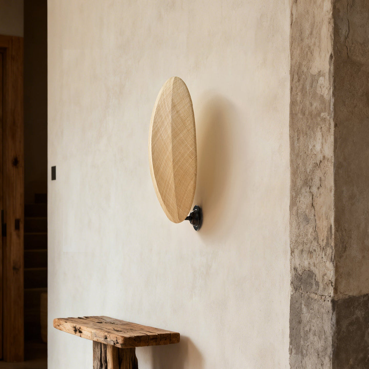 On a rough plaster wall above a rustic wooden bench, this Clérachic wall light shows its slim oval linen shade on a black mount.