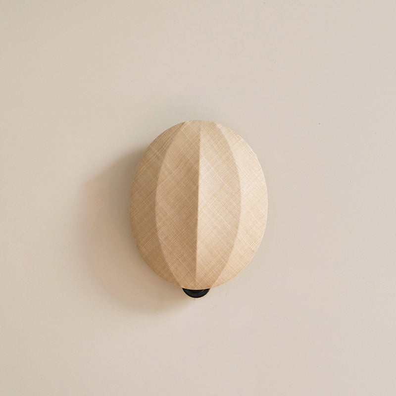 Close view of this Clérachic wall light on a plain cream wall, its rounded linen shade and its vertical ridge casting a soft shadow.