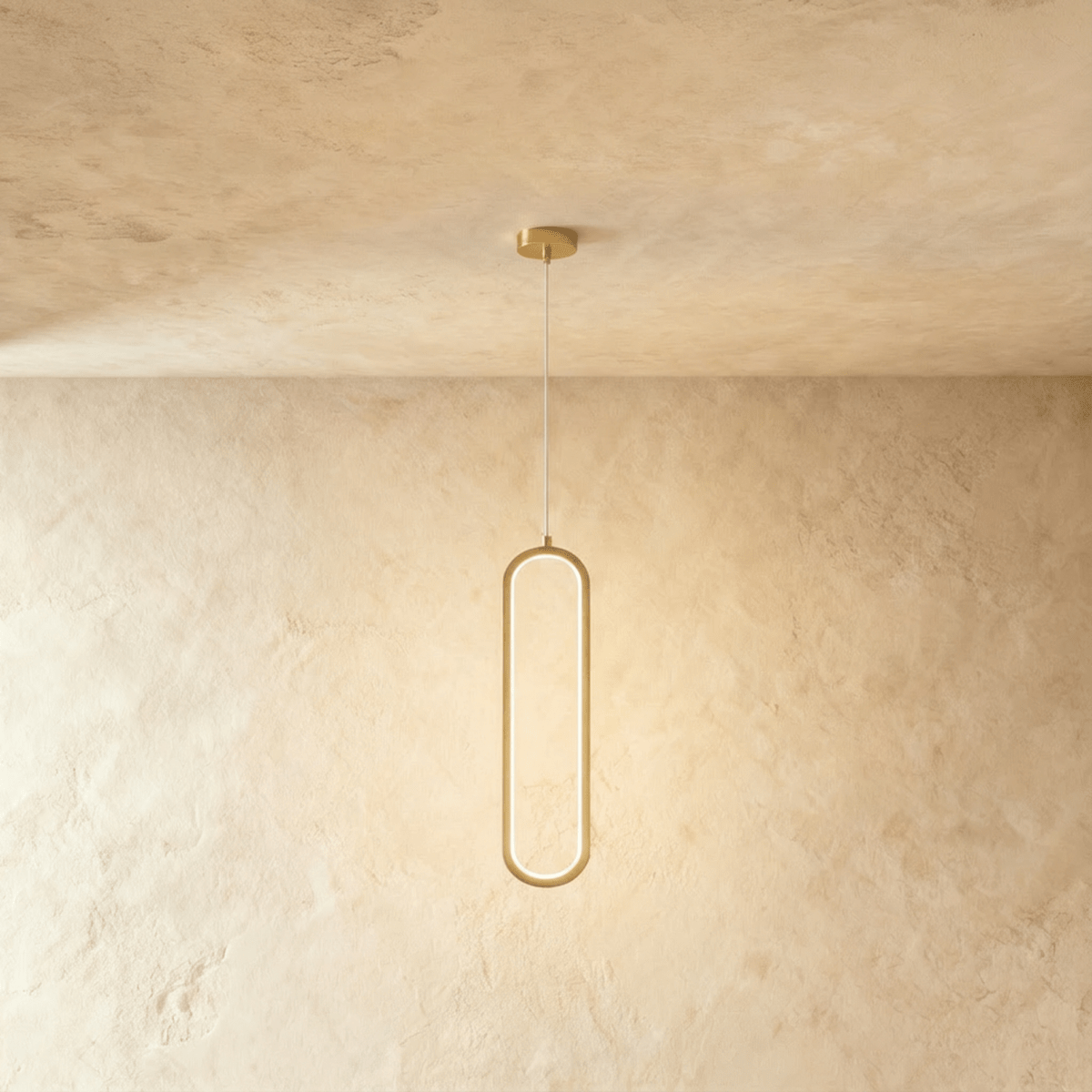 Clérachic slender brushed gold oval LED loop pendant glowing softly on a thin cord against a warm plaster wall.
