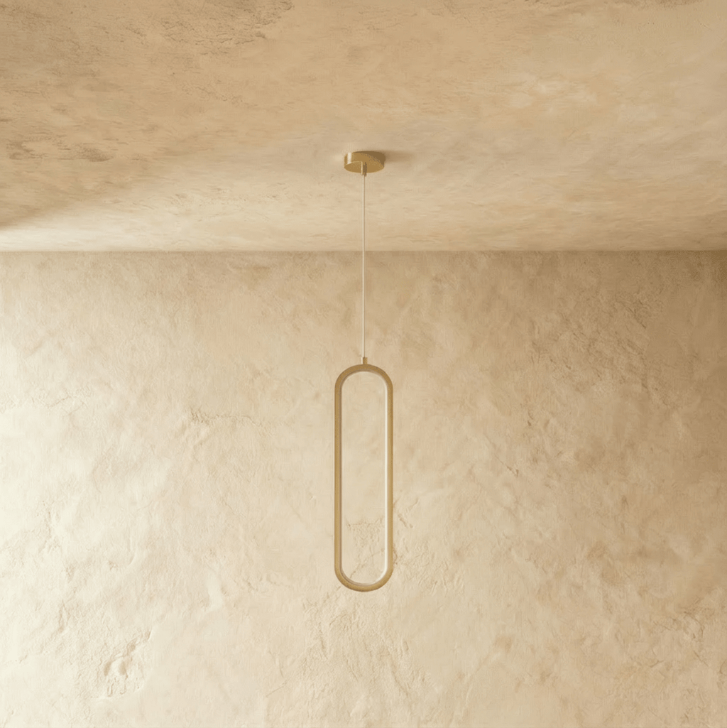 Clérachic slender brushed gold oval loop pendant, switched off, on a clear cord and gold canopy against plaster.