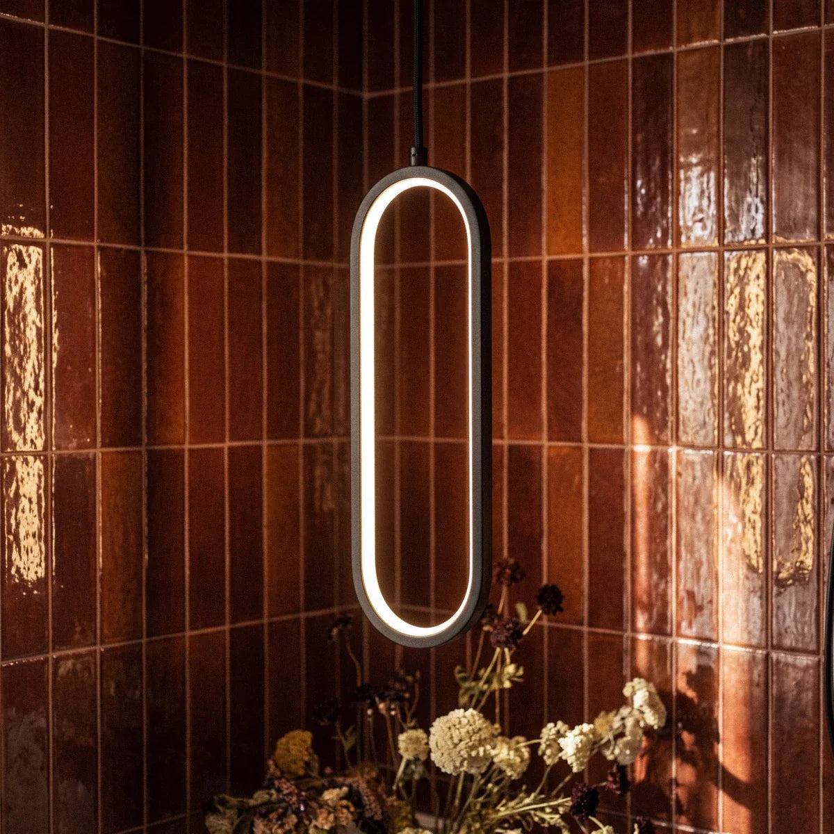 Close view of a Clérachic black oval LED loop pendant with a bright warm inner glow against glossy rust-red tiles.