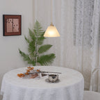 Clérachic pendant glowing beside sheer lace curtains, the round table and fern below catching its warm light.
