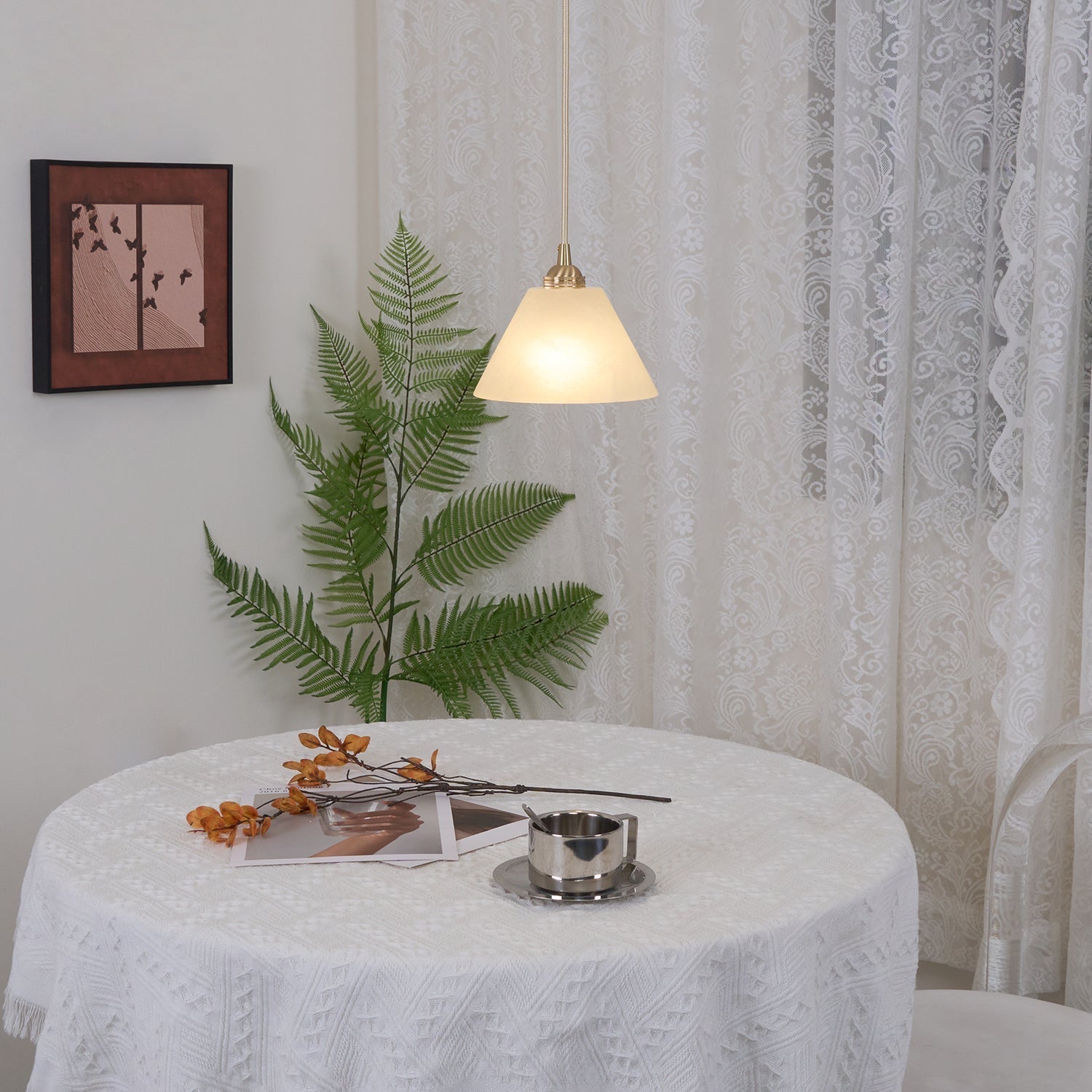 Clérachic pendant glowing beside sheer lace curtains, the round table and fern below catching its warm light.
