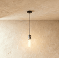 Clérachic marble pendant light hanging on a black cord against a warm plaster wall, its acrylic sleeve lit and casting a soft pool of light across the plaster.