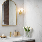 The Clérachic sconce lighting a marble bathroom wall, warm light spreading between the arched mirror and the stone vanity.