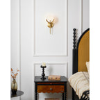 The Clérachic sconce on a white moulded panel above a dark spindle bedside table, its short brass stem tucked neat to the wall.