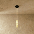 Clérachic slim alabaster cylinder pendant on a black cord and round black canopy, glowing against a limewashed wall.