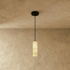Clérachic slim alabaster cylinder pendant on a black cord and round black canopy, glowing against a limewashed wall.