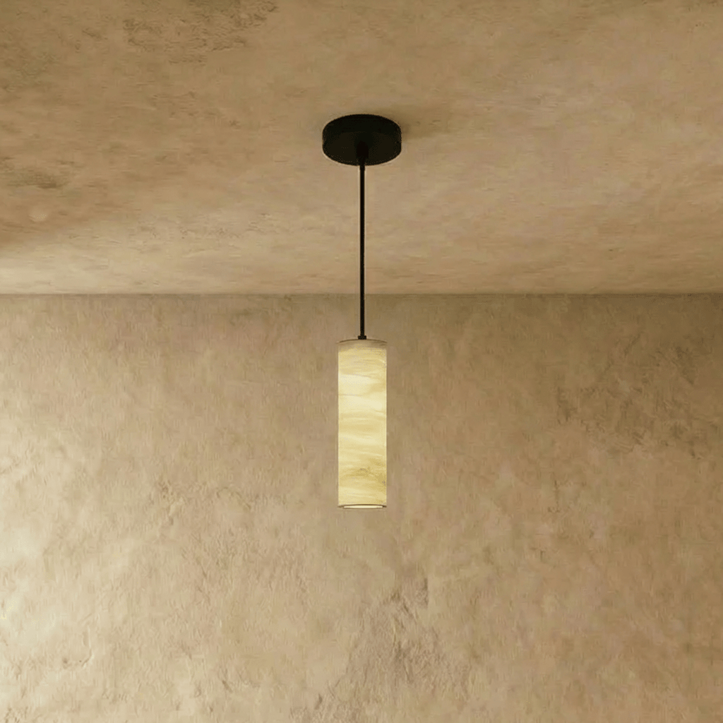 Clérachic alabaster cylinder pendant with a black rod and canopy, its veined stone softly lit against a textured plaster wall.