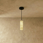 Clérachic alabaster cylinder pendant with a black rod and canopy, its veined stone softly lit against a textured plaster wall.