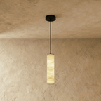 Clérachic taller alabaster cylinder pendant hung from a black cord and canopy, golden veining glowing on a plaster wall.