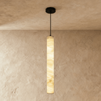 Clérachic tallest alabaster cylinder pendant on a black cord and canopy, its stone lit amber against a plaster wall.