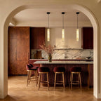 Row of three Clérachic alabaster cylinder pendants above a marble-topped kitchen island with velvet bar stools.