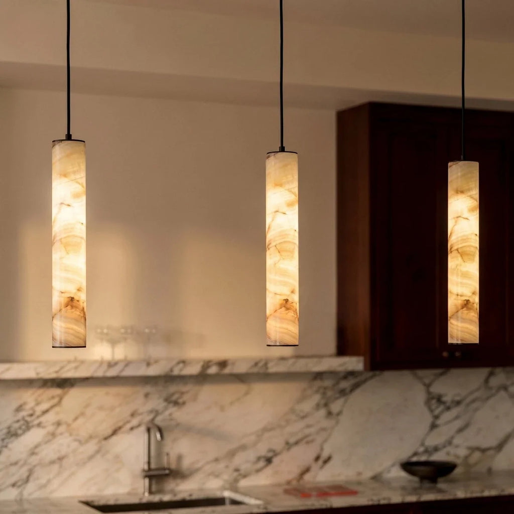 Three lit Clérachic alabaster cylinder pendants at different heights above a marble splashback, veining glowing amber.