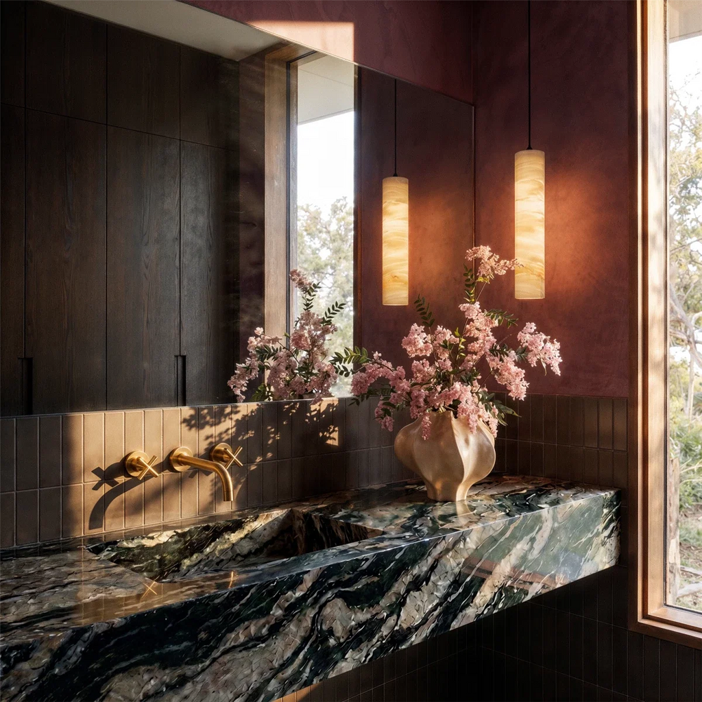 Two lit Clérachic alabaster cylinder pendants hung beside a mirror above a dark stone basin and brass taps.