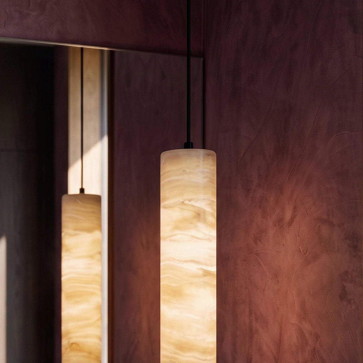 Close view of a lit Clérachic alabaster cylinder pendant, its layered amber veining reflected in a mirror beside it.