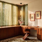 Clérachic alabaster cylinder pendant over a walnut corner desk with sheer green curtains and framed photographs.