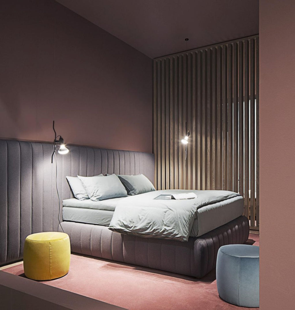 A Clérachic pendant spotlight lit above a low bed with a padded headboard, in a muted pink room with a yellow pouffe.