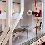 Red bendable-cord Clérachic pendant with a chrome spotlight head, in a white interior with large curved white pipes and a patterned bench.