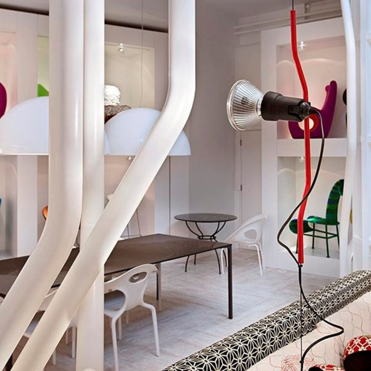 Red bendable-cord Clérachic pendant with a chrome spotlight head, in a white interior with large curved white pipes and a patterned bench.