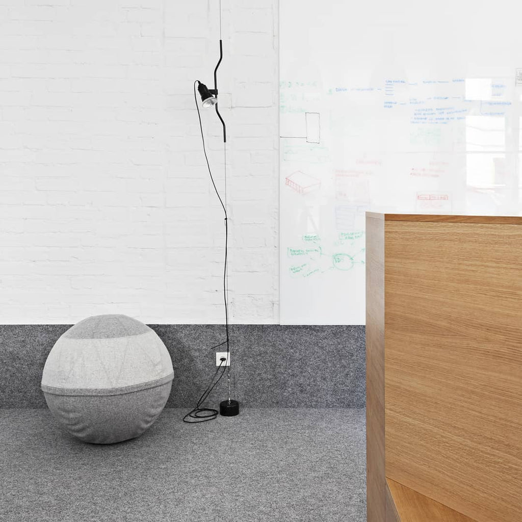 Clérachic black pendant spotlight on a long cable running down a white brick wall to a floor base, beside a grey ball chair.