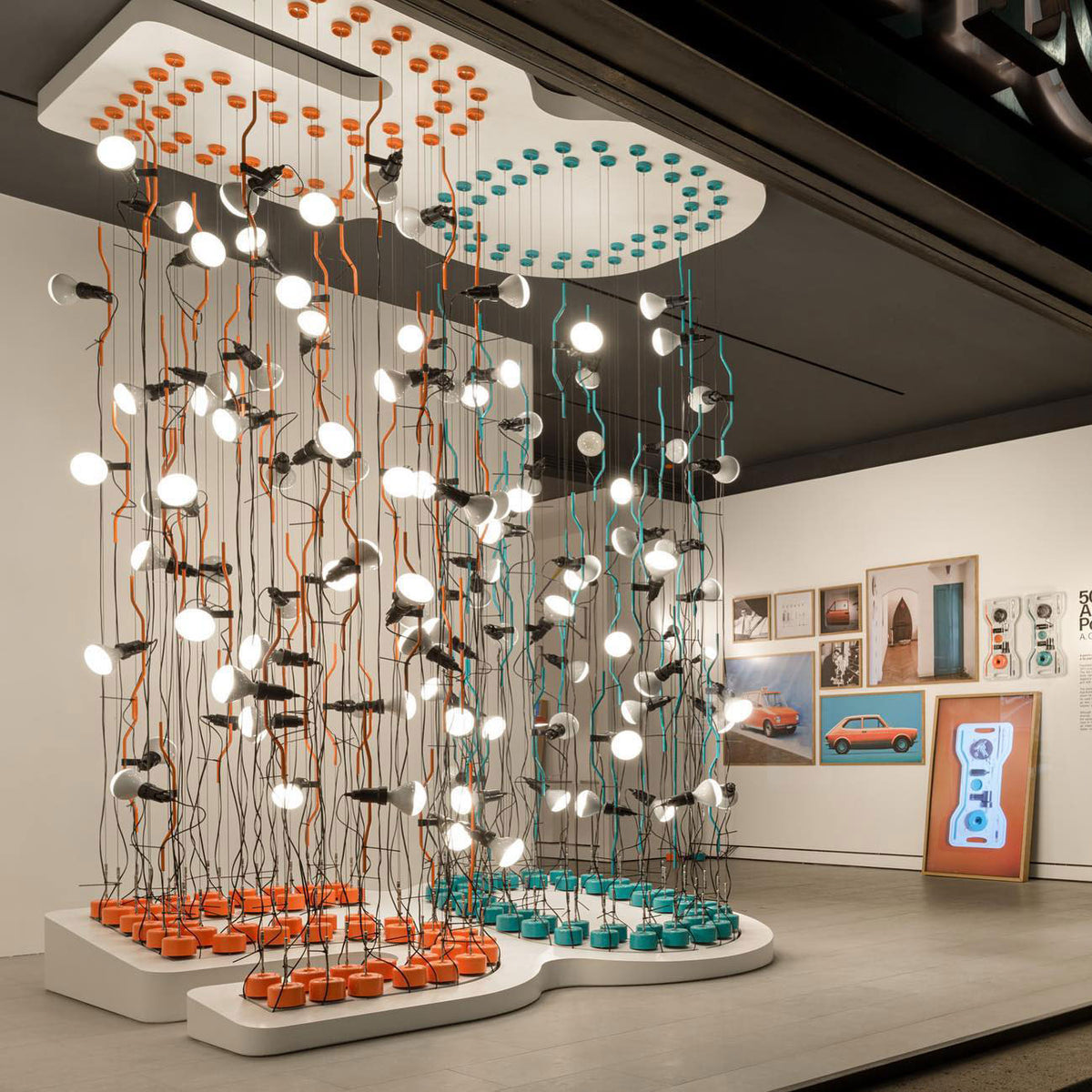 Large installation of lit Clérachic spotlight pendants hanging from orange and teal cords above round coloured bases in a gallery.